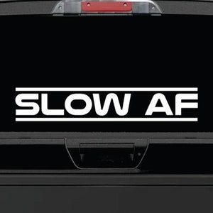 May include: A black truck with a white vinyl decal on the rear window that reads "SLOW AF".