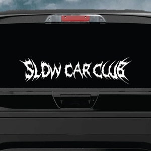May include: A white vinyl decal with the words "Slow Car Club" in a stylized metal font on a black truck tailgate.