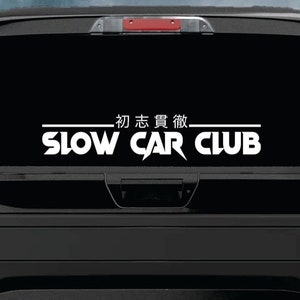 May include: A white vinyl decal with the text "Slow Car Club" in a stylised font. The text is in all caps and has a lightning bolt design in the "Car" portion. Above the text is a line with the Japanese characters "初 志 貫徹" in white.