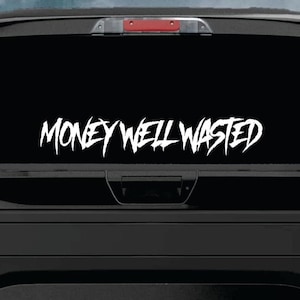 May include: White vinyl decal sticker with the text "Money Well Wasted" in a stylized font. The sticker is applied to the rear window of a black pickup truck.