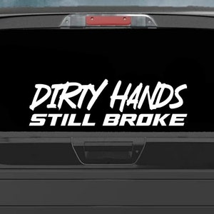 May include: A white vinyl decal with the text "DIRTY HANDS STILL BROKE" on a black truck tailgate.