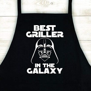 May include: Black apron with white text that reads "Best Griller in the Galaxy". A Darth Vader silhouette is above the text.