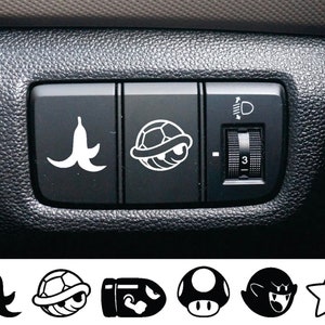 May include: A black car interior with a row of three buttons. The left button has a banana peel graphic, the middle button has a turtle shell graphic, and the right button has a knob with a light symbol and the number 3.
