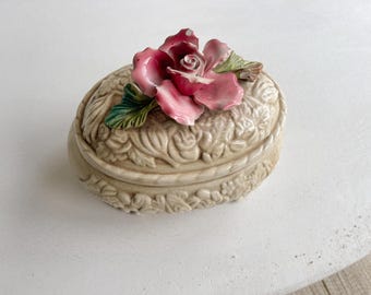 Puede incluir: Una caja de cerámica para baratijas con un diseño floral. La caja tiene forma ovalada y presenta una rosa rosa en relieve, tridimensional, con hojas verdes.