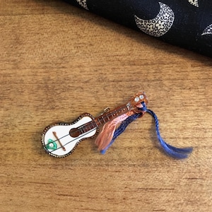 May include: A miniature ukulele pin with a white body, brown fretboard, and decorative trim. The pin has a small green and white design on the body and is accented with strands of pink and blue thread. The pin is on a wooden surface.