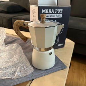 May include: A cream-coloured Moka Pot espresso maker with a wooden handle and lid knob. The pot is on a grey placemat, with the box behind it. The box reads "MOKA POT PRESSO 6 CUPS". The pot is designed to make 6 cups of espresso, or 300ml.