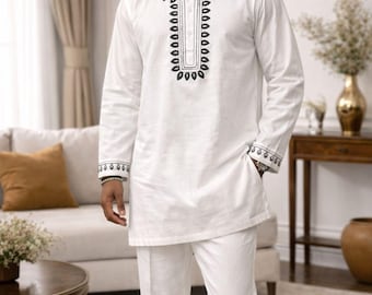 May include: A white long-sleeved tunic and matching trousers set featuring black embroidered patterns around the neckline, cuffs, and a coordinating white cap with black embroidery. The tunic has a buttoned placket and a straight hemline.
