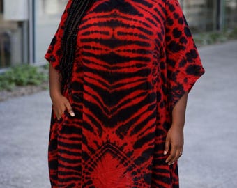 Puede incluir: Un vestido tipo caftán de corte holgado con un llamativo estampado tie-dye en rojo y negro. La prenda presenta un cuello redondo, mangas cortas y anchas, y bolsillos laterales visibles. La tela muestra un diseño abstracto y simétrico.