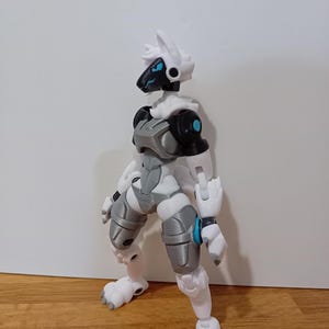 May include: An articulated action figure in white and grey with a robotic design. The figure has a black head with blue accents, a white helmet, and grey and white armour. The figure is posed standing with its feet in a paw-like shape.