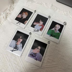 May include: Five rectangular instant film photographs featuring portraits of individuals. Each photo has a white border, a handwritten number in the top right corner, and a black ink signature at the bottom. The subjects wear various coloured shirts.
