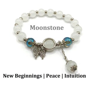 May include: White moonstone beaded bracelet with a silver butterfly charm and a dangling moonstone bead. The bracelet has a silver clasp and two iridescent blue beads. The text "Moonstone" is written on the image. The text "New Beginnings | Peace | Intuition" is written below the bracelet.