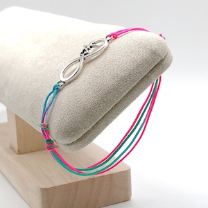 May include: A friendship bracelet with an infinity symbol charm. The bracelet is made with three strands of cord in pink, green, and purple.