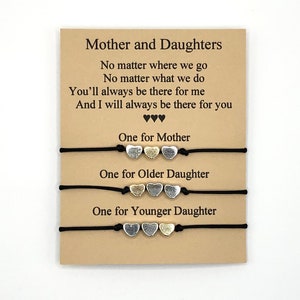 Puede incluir: Un conjunto de tres pulseras a juego con dijes en forma de corazón, presentadas en una tarjeta con el texto "Mother and Daughters". Cada pulsera tiene tres dijes de corazón en tonos plateados y dorados sobre un cordón negro.