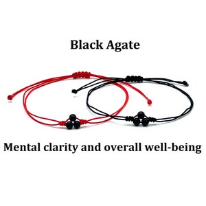 Op de afbeelding: Twee rode en zwarte armbandjes van touw met zwarte agaat kralen. De tekst "Black Agate" en "Mental clarity and overall well-being" is zichtbaar.