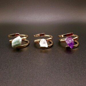 May include: Three gold rings with different colored gemstones. The rings are made of wire wrapped around a stone. The stones are green, white, and purple.