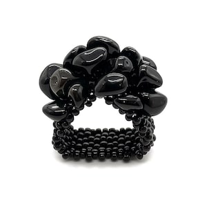 May include: A black beaded ring with a cluster of polished black stones. The ring features a wide band made of small, round black beads. The stones are irregularly shaped and add a unique texture to the ring.