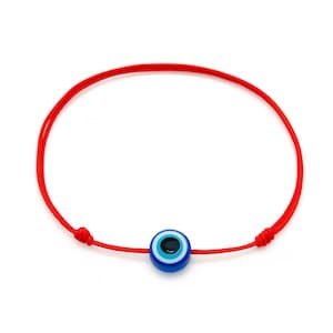 May include: A red string bracelet featuring a blue and white evil eye charm. The charm has a black center and is surrounded by concentric circles of blue and white. The bracelet is tied with knots for adjustment.