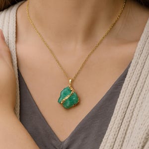 K&ouml;nnte beinhalten: Eine goldene Halskette mit einem rohen, gr&uuml;nen Edelsteinanh&auml;nger. Der Anh&auml;nger hat eine unregelm&auml;&szlig;ige Form und ist mit einem d&uuml;nnen Goldband umwickelt. Ein passendes goldenes Armband mit einem rechteckigen gr&uuml;nen Stein ist ebenfalls sichtbar.