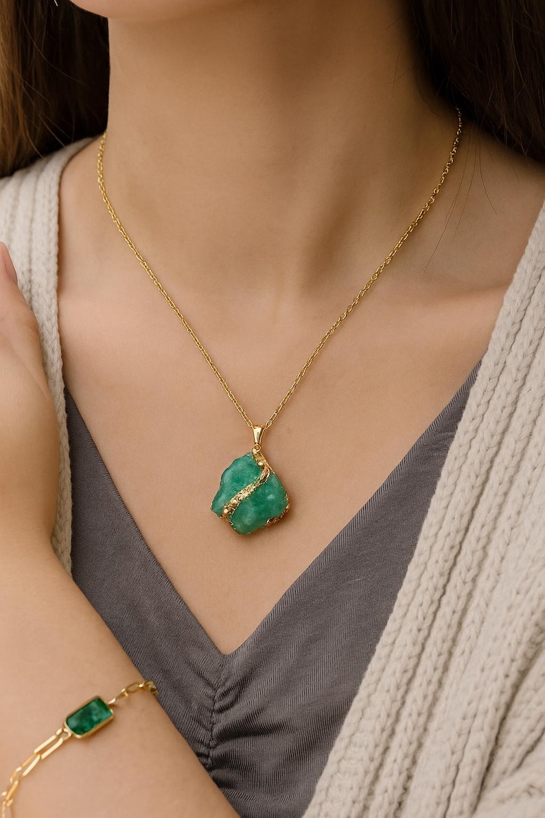 K&ouml;nnte beinhalten: Eine goldene Halskette mit einem rohen, gr&uuml;nen Edelsteinanh&auml;nger. Der Anh&auml;nger hat eine unregelm&auml;&szlig;ige Form und ist mit einem d&uuml;nnen Goldband umwickelt. Ein passendes goldenes Armband mit einem rechteckigen gr&uuml;nen Stein ist ebenfalls sichtbar.