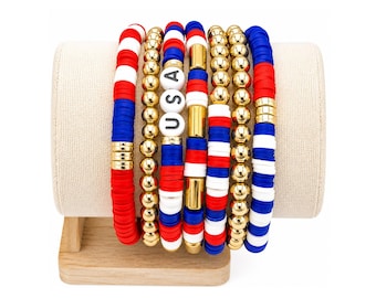 May include: A set of six stacked bracelets featuring red, white, and blue polymer clay discs, metallic gold beads, and cylindrical gold accents. One bracelet includes white letter beads spelling out USA in black font, arranged vertically on the display.