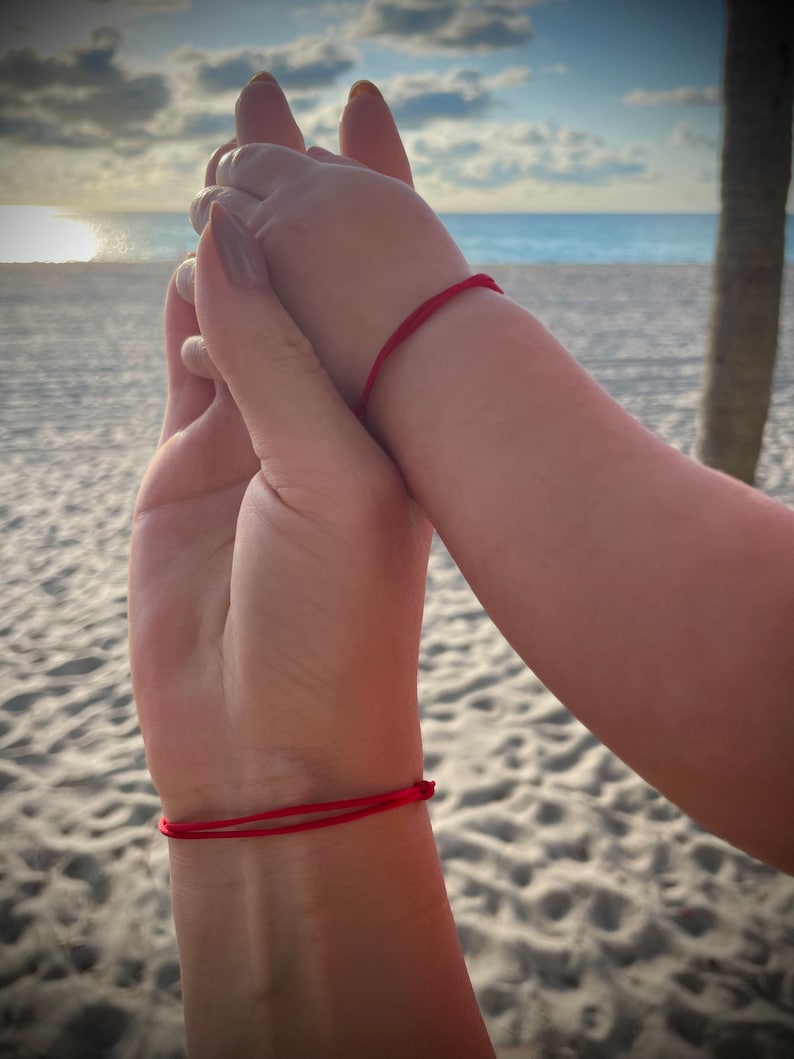 May include: Two people holding hands with red string bracelets on their wrists. The image is taken on a beach with white sand and a blue sea in the background.
