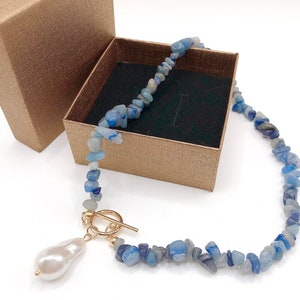 May include: A blue gemstone necklace with a large white pearl pendant and gold-colored clasp. The necklace is displayed inside a small, open, textured, brown gift box. The gemstones are small, irregular chips of blue and white.