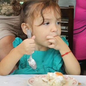 May include: A young girl with brown hair in pigtails is eating a piece of cake with a spoon. She is wearing a turquoise dress and has a red bracelet on her wrist. The cake is on a white plate with a pink and white design.