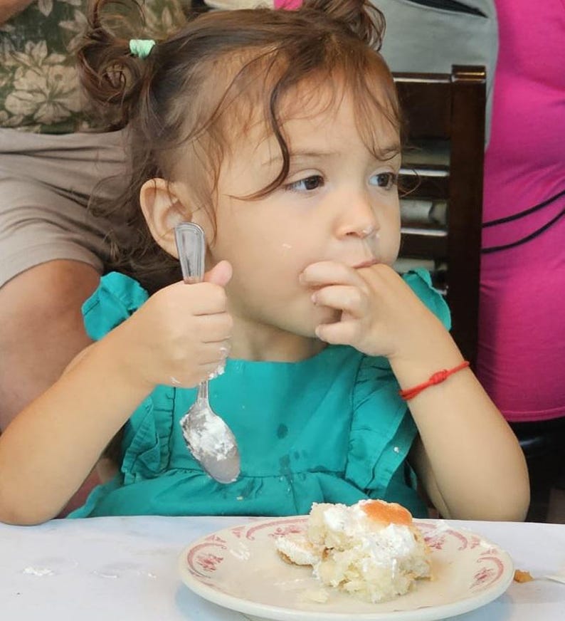 May include: A young girl with brown hair in pigtails is eating a piece of cake with a spoon. She is wearing a turquoise dress and has a red bracelet on her wrist. The cake is on a white plate with a pink and white design.