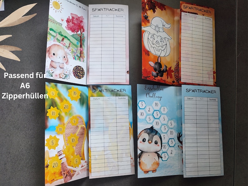 May include: Four rectangular paper savings trackers with colourful illustrations and grids labelled SPARTRACKER with columns for Datum, +/-, and Summe. Designs include a bunny, a pumpkin, sunflowers, and a penguin. Text reads Passend f&uuml;r A6 Zipperh&uuml;llen.