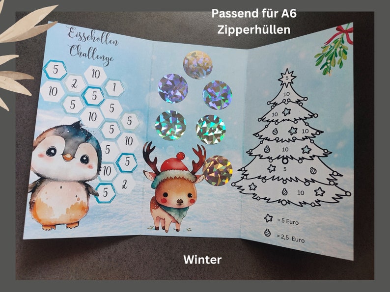 May include: A tri-fold savings challenge card featuring a penguin, a reindeer, and a Christmas tree. The card includes hexagonal fields with numbers, holographic circular stickers, and text reading Eisschollen Challenge, Winter, and Passend f&uuml;r A6 Zipperh&uuml;llen.