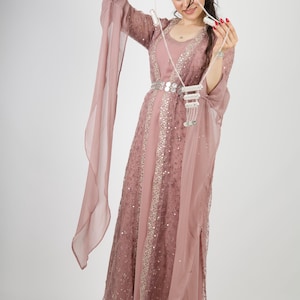 Könnte beinhalten: Ein bodenlanges Kleid in Altrosa mit einem transparenten Überwurf, der mit aufwendigen silbernen Pailletten- und Perlenstickereien verziert ist. Das Kleid hat lange, fließende Ärmel und einen passenden Gürtel mit silbernen Akzenten sowie eine lange Kette.