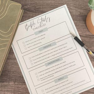 May include: A Bible study checklist with sections for Pray, Observe, Understand, and Apply. A pen rests on the checklist, next to a green and gold journal and a small potted plant. The checklist is on a wooden surface.