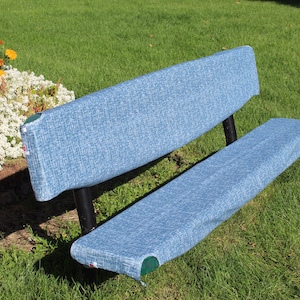 May include: A blue bench with a textured fabric seat and backrest. The bench has black metal legs and is situated on a grassy lawn, with flowers in the background. The bench is designed for outdoor use.
