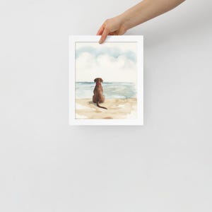 May include: Watercolor painting of a brown dog sitting on a beach, looking out at the ocean. The sky is blue with white clouds.