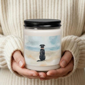 May include: A white candle jar with a black lid, featuring a watercolor illustration of a black dog with an orange collar sitting on a beach, gazing at the ocean. The candle is held in a person's hands.