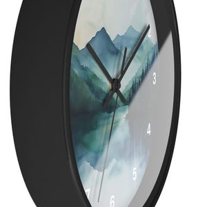 May include: A round black wall clock with a watercolor landscape design. The clock face features a mountain and lake scene in shades of blue and green. The clock hands are black, and the numbers are white.