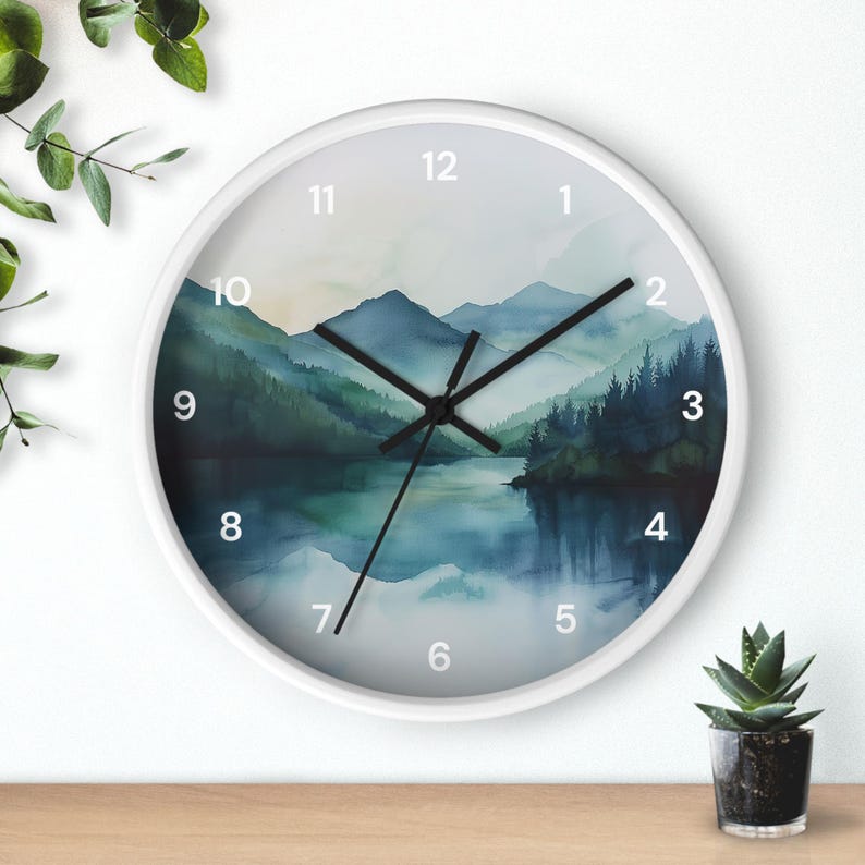 May include: White-framed wall clock with a watercolor landscape design. The clock face features a serene scene of mountains, a lake, and trees in shades of blue and green. Black clock hands and white numerals complete the design.