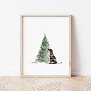 May include: Watercolor print of a Christmas tree with a star on top, adorned with lights. A brown and white dog sits beside the tree. The artwork is framed in a light wood frame.