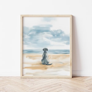 May include: Watercolor print of a dog sitting on a beach, facing the ocean. The artwork features a light blue dog with a blue collar, sandy beach, and a cloudy sky. The framed print is displayed on a white wall.