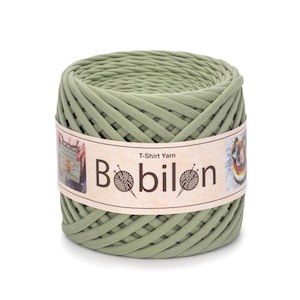 Op de afbeelding: Een cilindrische spoel van saliegroen T-shirtgaren. Het garen is strak opgerold en omwikkeld met een label met de tekst "Bobilon" met een logo van garen en naalden. Op het label staat ook "T-Shirt Yarn".