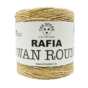 Può includere: Una bobina di rafia color naturale. L'etichetta recita "RAFFIA SWAN ROUND" con il marchio "TRE SFERE" e l'indirizzo del sito web. Lo spago è ben avvolto e l'aspetto generale suggerisce un articolo per l'artigianato o il giardinaggio.