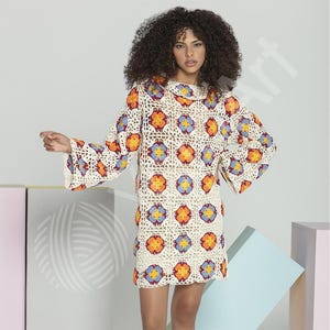 May include: A cream-colored crochet dress with a floral pattern in orange, yellow, and blue. The dress has long, flared sleeves and a round neckline. The model is wearing the dress.