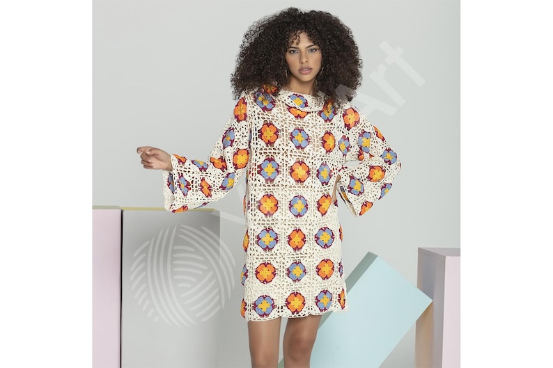 May include: A cream-colored crochet dress with a floral pattern in orange, yellow, and blue. The dress has long, flared sleeves and a round neckline. The model is wearing the dress.