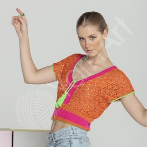 May include: Orange crochet top with short sleeves, a V-neck, and a pink and green trim. The top has a tassel detail at the neckline. The model is wearing blue jeans. The top is a summer fashion item.