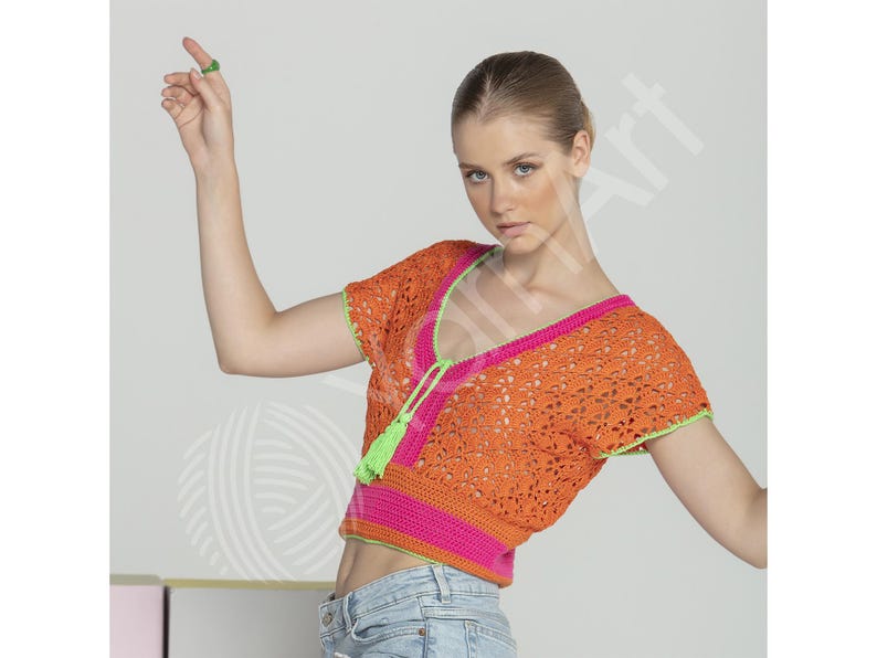 May include: Orange crochet top with short sleeves, a V-neck, and a pink and green trim. The top has a tassel detail at the neckline. The model is wearing blue jeans. The top is a summer fashion item.