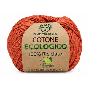 Puede incluir: Un ovillo de hilo de algodón color naranja quemado envuelto en una etiqueta de papel color canela. La etiqueta muestra el texto TRE SFERE, FILATI TRE SFERE, COTONE ECOLOGICO, 100% Riciclato, Made in Italy, un icono de hoja verde y banderas.