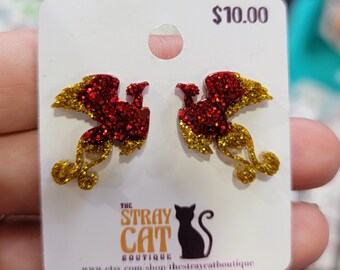 May include: A pair of red and gold glitter phoenix earrings on a white card. The earrings have a detailed design with a red body and gold wings. The card has the text "The Stray Cat Boutique" and a black cat silhouette.