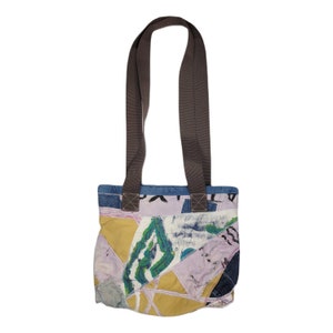 Puede incluir: Una bolsa de mano de patchwork con asas marrones. La bolsa está hecha de diferentes telas en tonos de azul, rosa, amarillo y verde. La tela tiene una variedad de patrones, incluyendo florales, geométricos y abstractos.