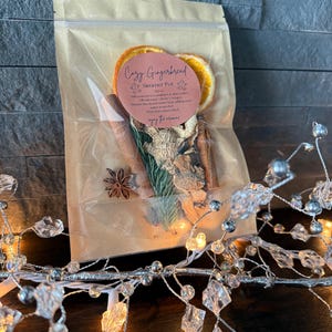 May include: A clear plastic pouch containing dried orange slices, cinnamon sticks, star anise, ginger pieces, and pine needles. A round pink label on the front reads Cozy Gingerbread Simmer Pot with instructions and the phrase enjoy the aroma.