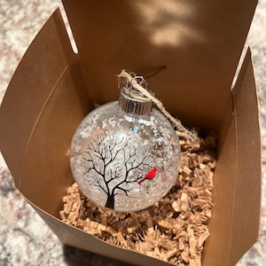 May include: A clear glass spherical ornament featuring a black silhouette of a bare tree and a small red cardinal bird perched on a branch. The interior is filled with white glitter, and the ornament hangs from a brown twine string inside a box.
